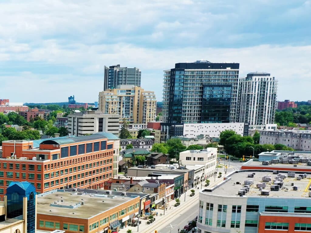 A cityscape with mid- and high-rise buildings under a partly cloudy sky, viewed from above a busy street lined with smaller shops and offices.