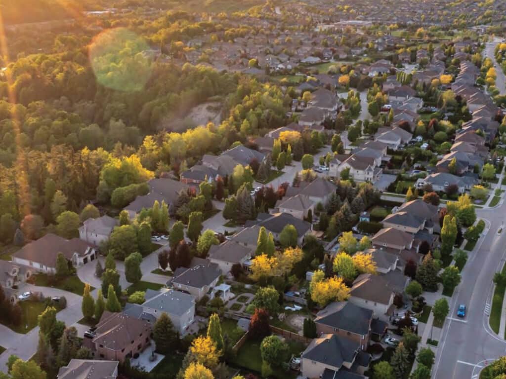Aerial view of a suburban neighbourhood at sunset, showing tree-lined streets, houses, and roads with a mix of greenery and built environment.