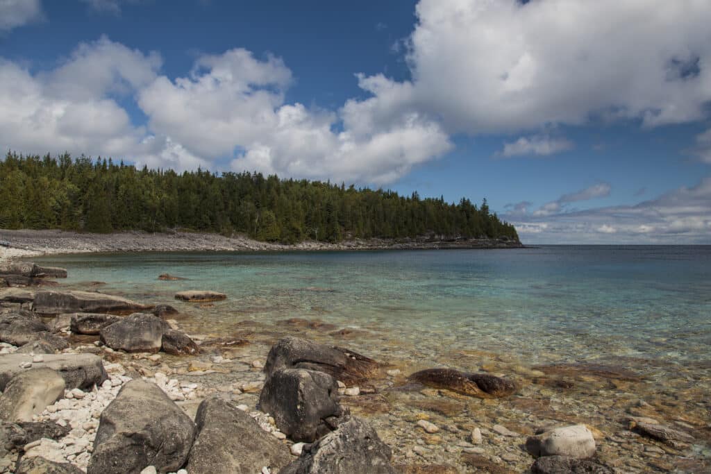 View of a beach with bright blue water, blue sky with clouds and a rocky shore.
