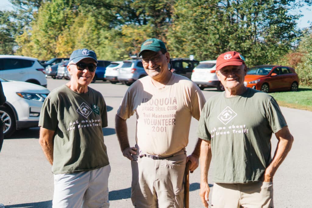 Three smiling men wearing hats and t-shirts stand in a parking lot.