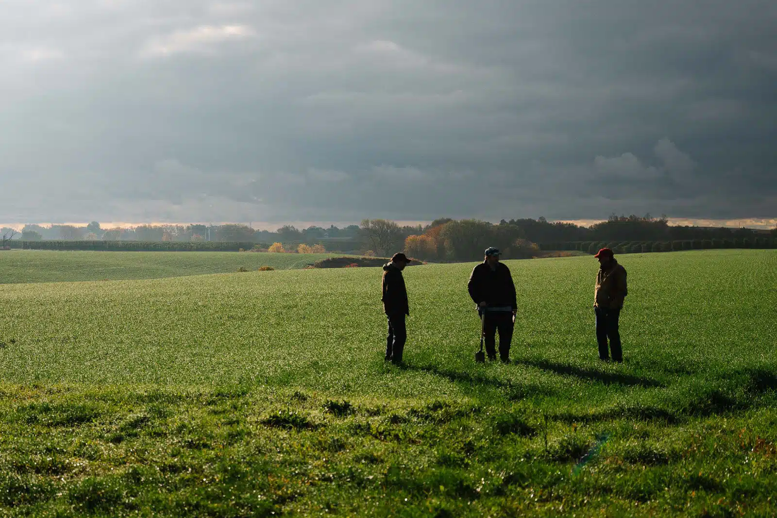 Three people talking in a farm field with a distant line of autumn-colored trees.