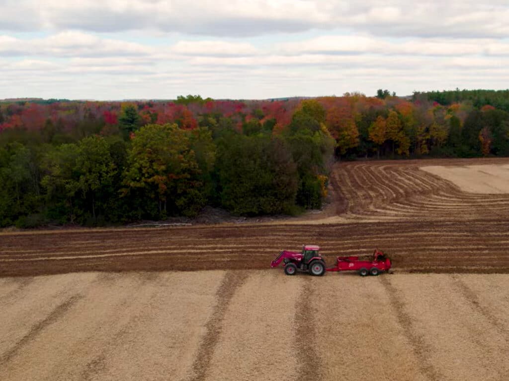 Aerial view of a farm field in autumn, with a red tractor pulling equipment across harvested rows beside a forest of colourful fall trees under a cloudy sky.